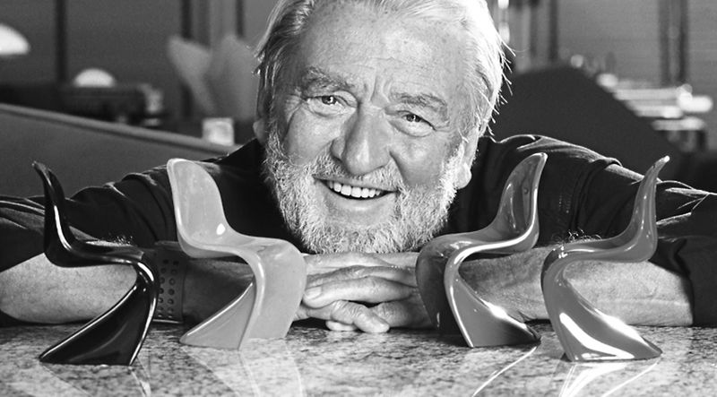 Verner Panton portrait en noir et blanc. Il sourit et a une moustache ainsi qu'une barbe. Il est accoud&eacute; sur une table sur laquelle est pos&eacute;e quatre miniatures de chaises S.
